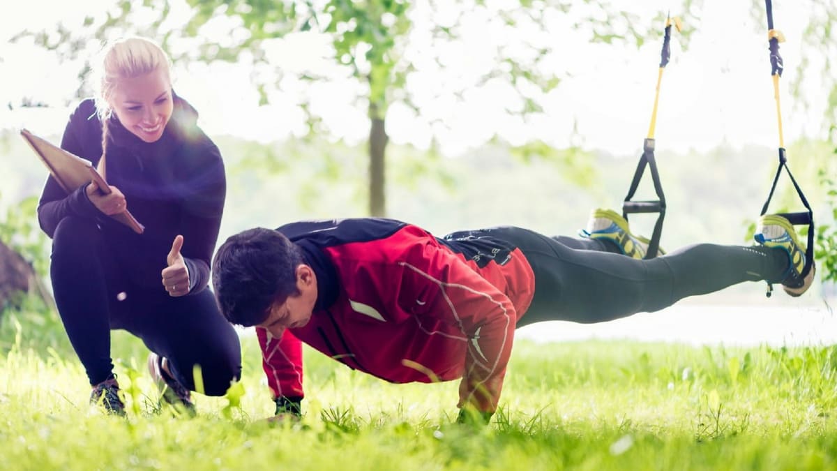 Personal trainer agachada na grama, com prancheta, faz sinal de positivo para aluno em flexão com os pés em fitas suspensas