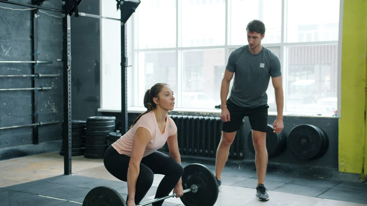 Aluna se agacha para levantar uma barra no chão enquanto o personal trainer observa de pé