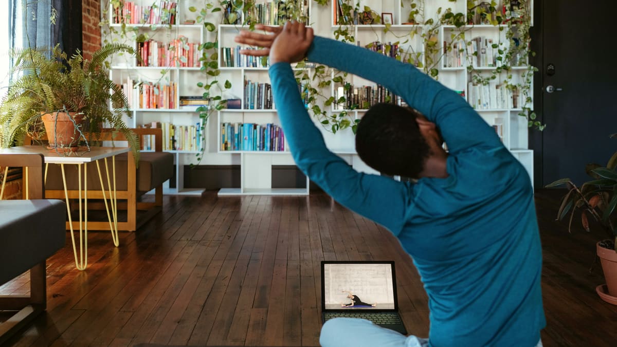 Homem de costas, sentado em um tapete, alonga o tronco acompanhando uma aula exibida em um notebook no chão