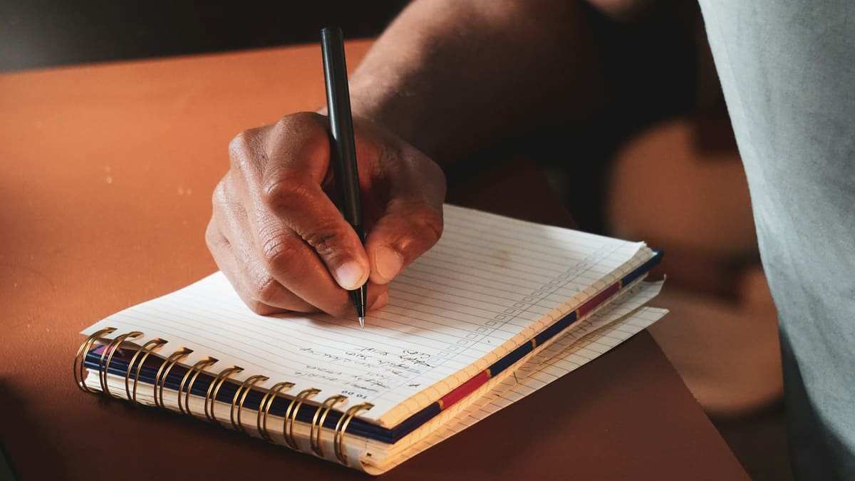Mão anota uma lista à caneta em um caderno espiral apoiado sobre uma mesa