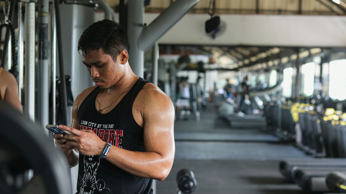 Homem suado em uma academia olha mensagens no celular, com fileiras de esteiras desfocadas ao fundo